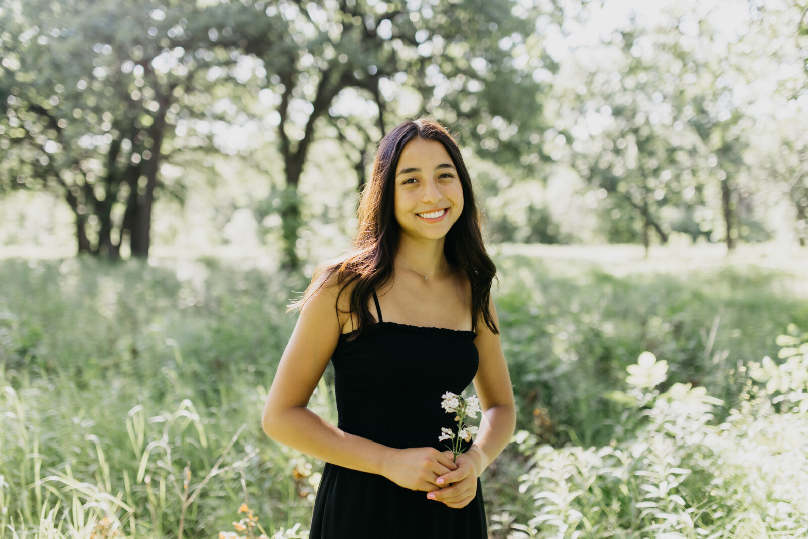 A smiling high school senior in her black dress captured against a backdrop of nature in a photograph.