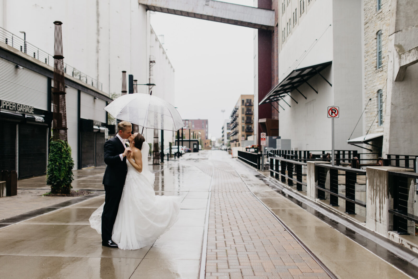 A photo of a newlywed couple kissing on the streets while carrying a clear dome umbrella