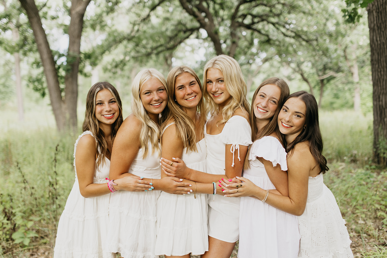 a photo of high school seniors wearing white dresses on a field with trees