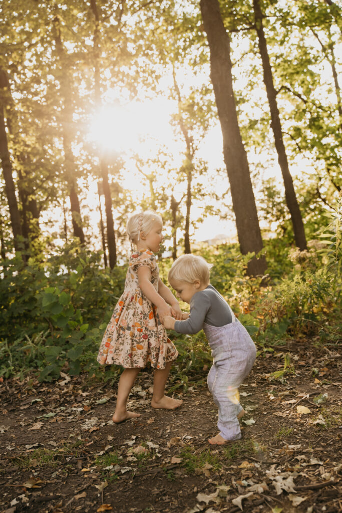 Two young siblings holding hands and playing barefoot in the woods during golden hour in Minnesota.