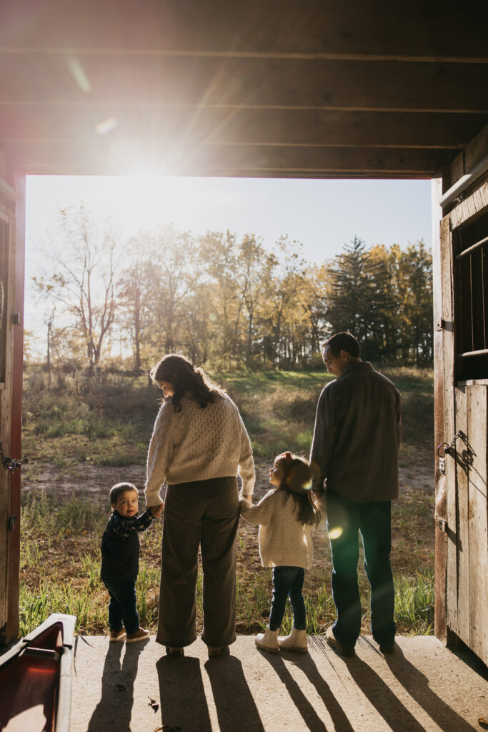 Family of four standing in the doorway of a barn, holding hands and looking out at a sunlit Minnesota field during a fall family photo session.