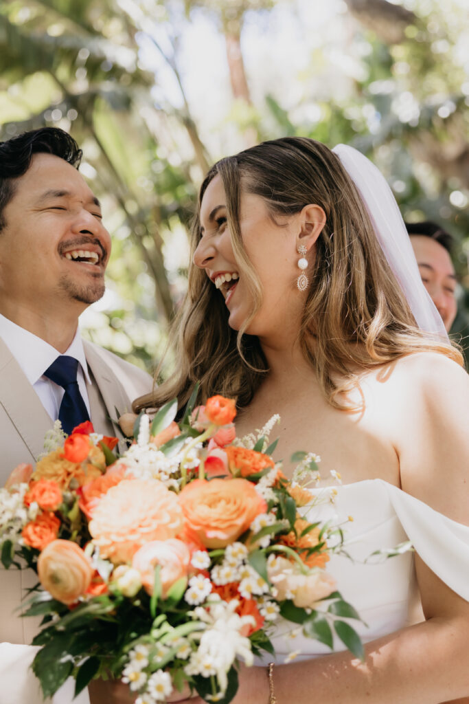 Bride and groom laughing together while holding a vibrant orange and coral bouquet during their Montecito private estate wedding.