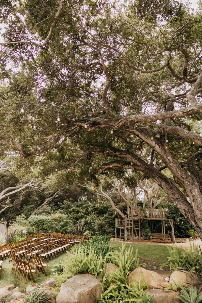 Outdoor wedding ceremony setup at a Montecito private estate beneath oak trees with string lights and wooden chairs.