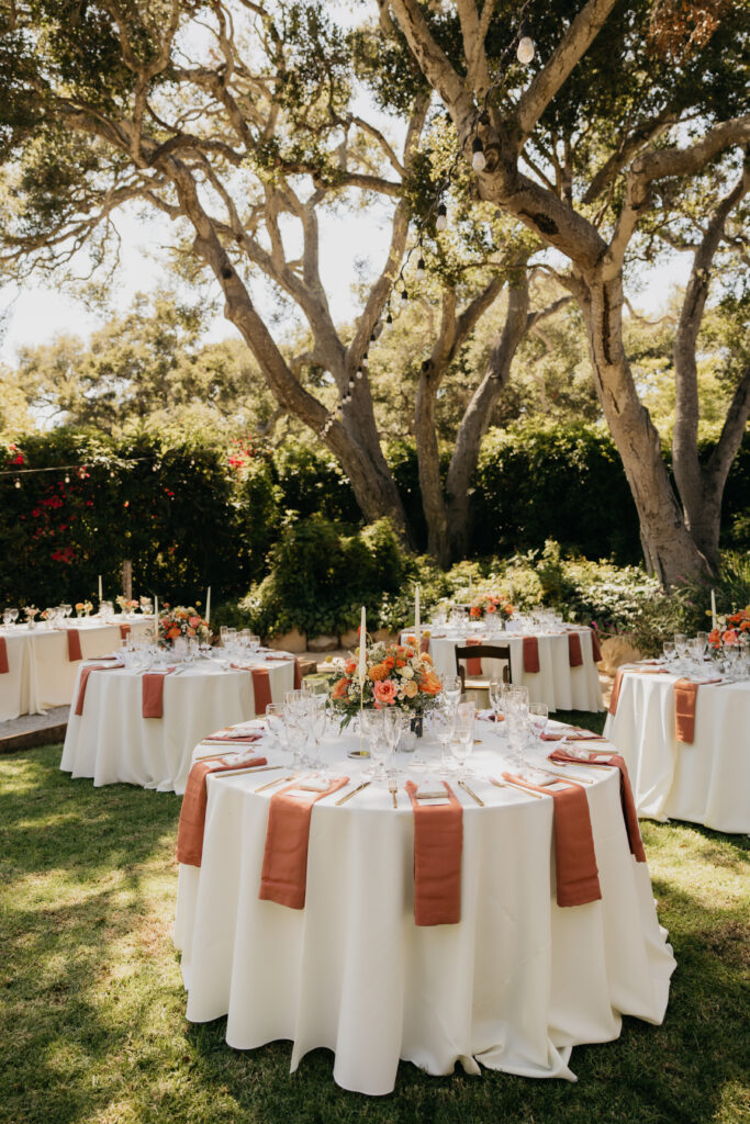 Wedding reception tables with terracotta napkins and floral centerpieces set on the lawn of a Montecito private estate.