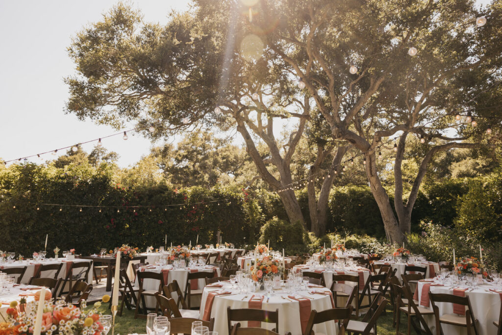 Outdoor wedding reception tables set beneath oak trees with terracotta napkins and floral centerpieces in Montecito.