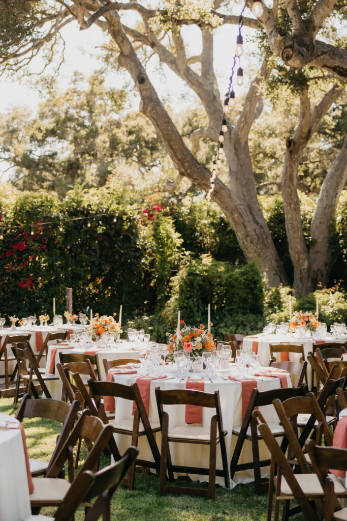Outdoor wedding reception tables with terracotta napkins and floral centerpieces set beneath oak trees at a Montecito private estate.