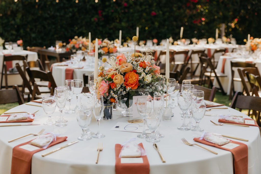 Close-up of a lush orange and coral floral centerpiece with taper candles on a wedding reception table in Montecito.