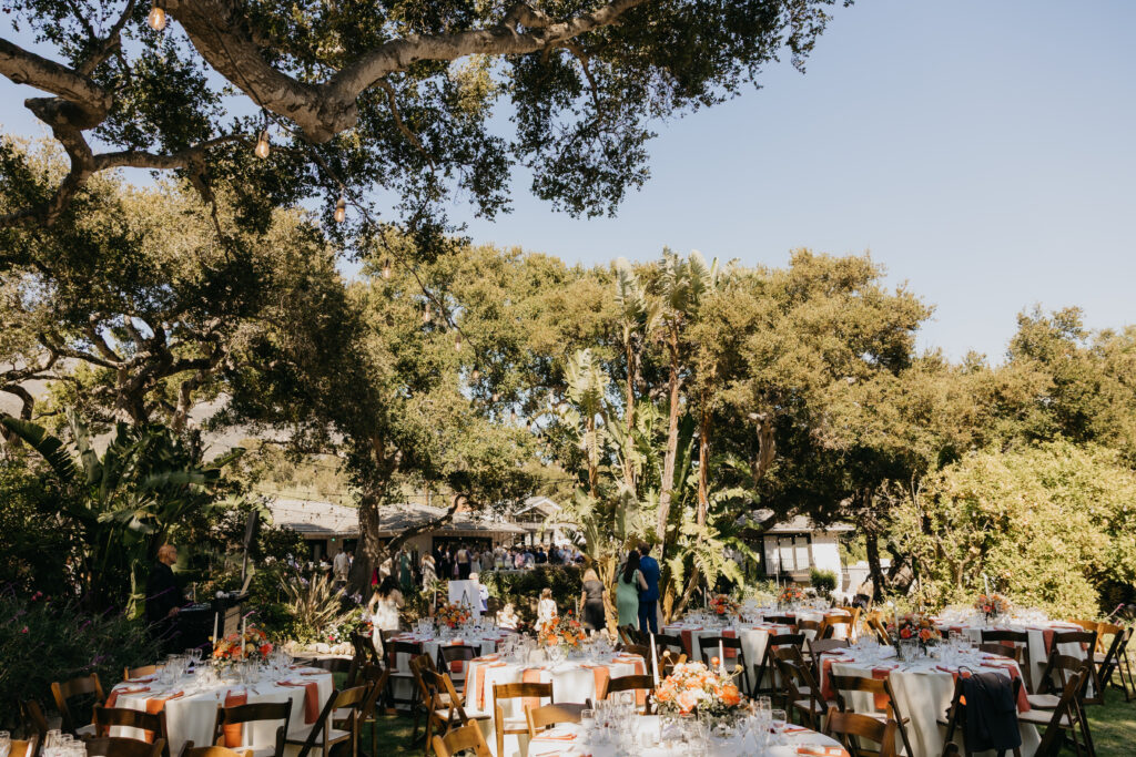 Outdoor wedding reception setup at a Montecito private estate with round tables, floral centerpieces, and oak trees overhead.