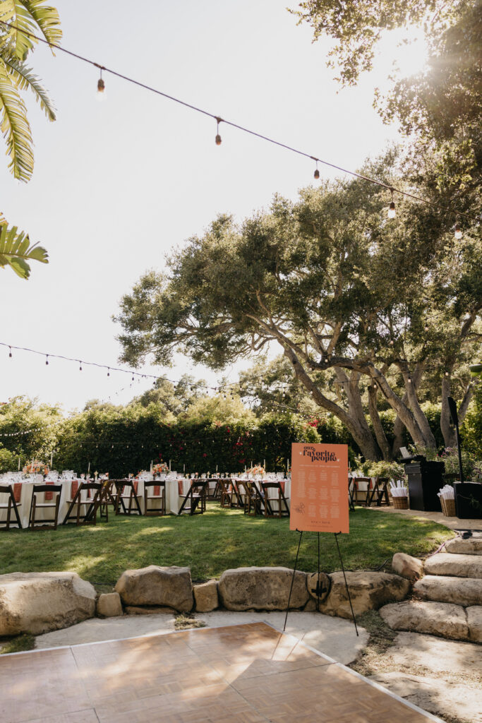 Wedding reception seating chart displayed on an orange sign with wooden chairs and string lights at a Montecito estate.