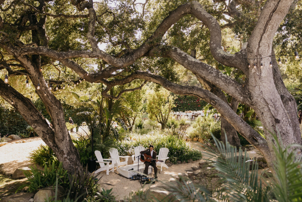 Ceremony musician playing acoustic guitar beneath oak trees at a Montecito private estate wedding.