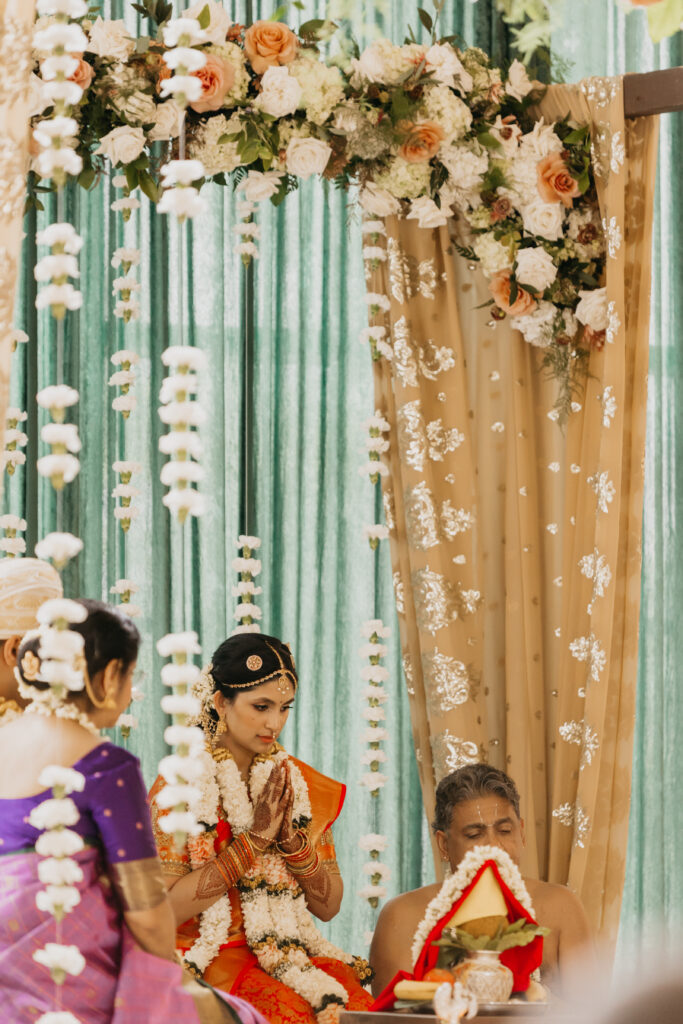 Bride Apoorva seated with family, hands folded in prayer during Indian wedding rituals beneath mandap