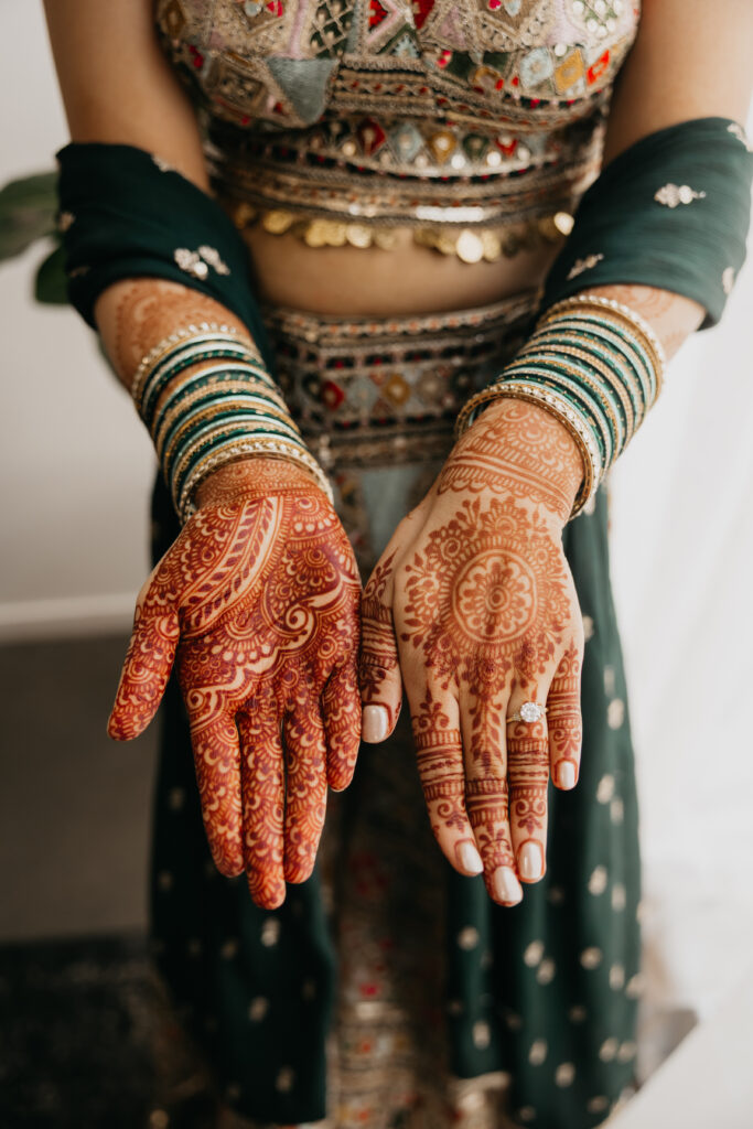 Close-up of bride’s hands adorned with intricate mehndi designs and green glass bangles, with part of her embroidered lehenga and dupatta visible in the background