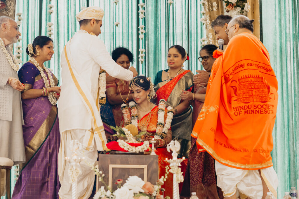 Groom Sanjay places sacred ornament on bride Apoorva’s head during Indian wedding ceremony surrounded by family and priest at Renaissance Depot Minneapolis | Mycah Bain Photography