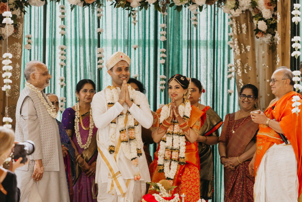 Newlyweds Apoorva and Sanjay standing together with folded hands, adorned in garlands during sacred Indian wedding rituals | Mycah Bain Photography