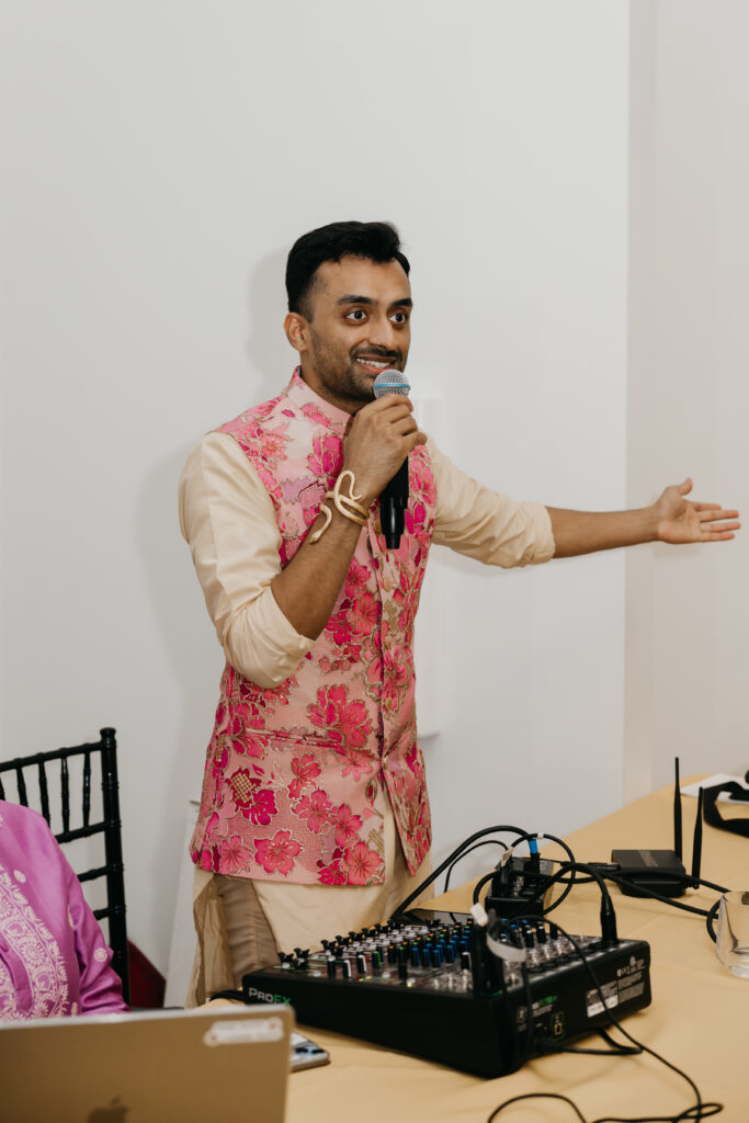 Man in a pink floral embroidered vest speaking into a microphone at a sangeet in Minneapolis, standing behind a sound mixer and smiling as he addresses guests