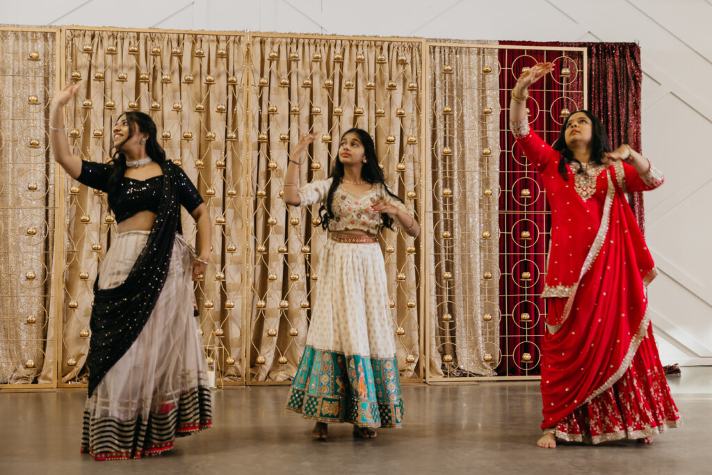 Three women dancing on stage during a sangeet in Minneapolis, dressed in traditional Indian lehengas with a golden and red curtain backdrop