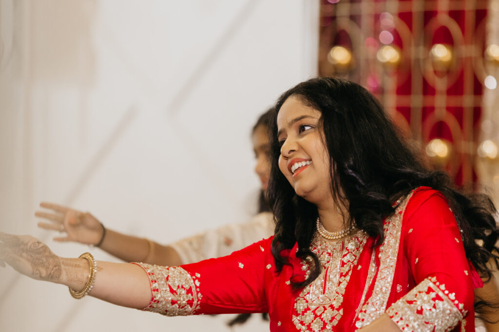 Woman in a red and gold embroidered outfit smiling and dancing during a sangeet celebration, with mehndi on her hands and joy in her expression