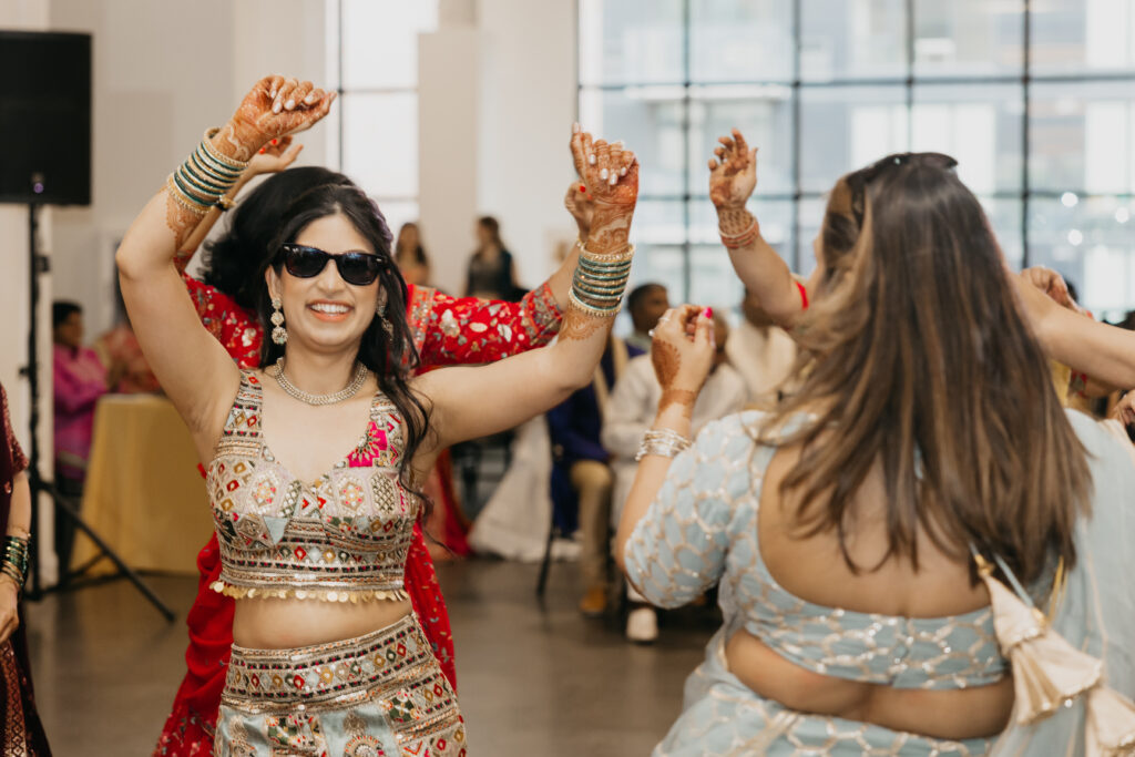 Bride dancing with sunglasses on during the sangeet, surrounded by guests in colorful lehengas, with mehndi on her hands and a big smile on her face