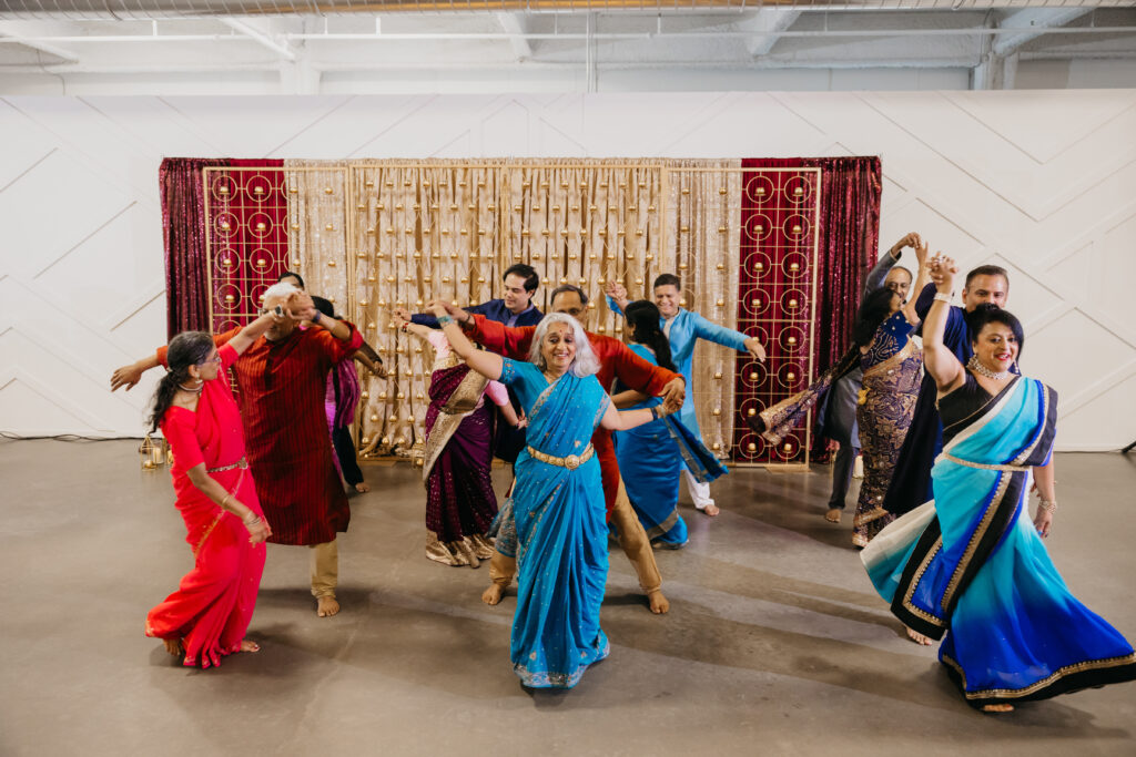 A group of wedding guests performing a joyful group dance during a sangeet in Minneapolis, dressed in colorful traditional Indian attire including bright saris and kurtas, with a gold and maroon backdrop behind them