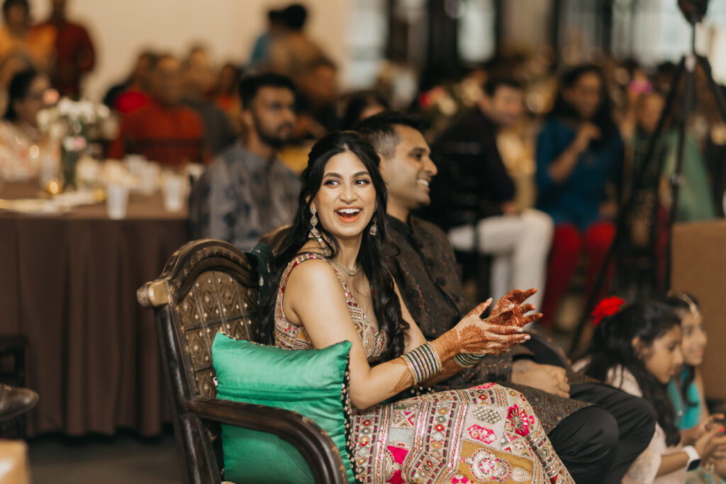Bride laughing during the sangeet celebration, seated beside the groom, wearing a multicolored embroidered lehenga with green bangles and mehndi on her hands