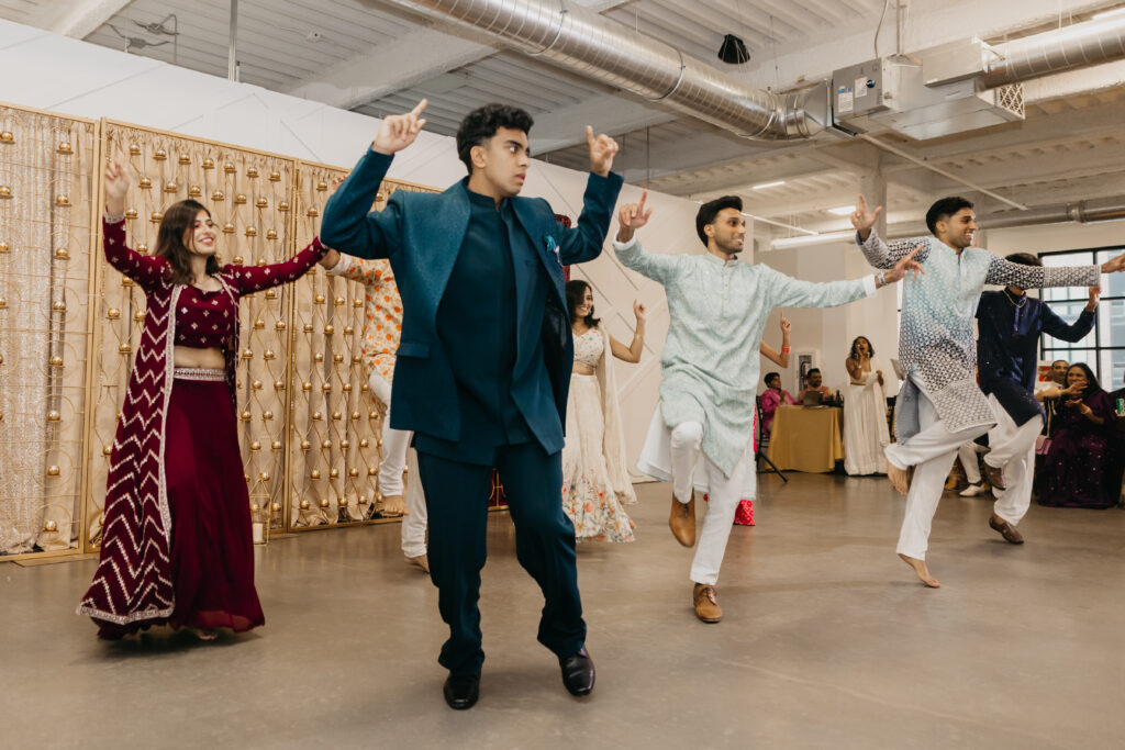 A group of wedding guests performing a choreographed dance during the sangeet, wearing coordinated outfits in jewel tones and pastels in front of a gold curtain backdrop
