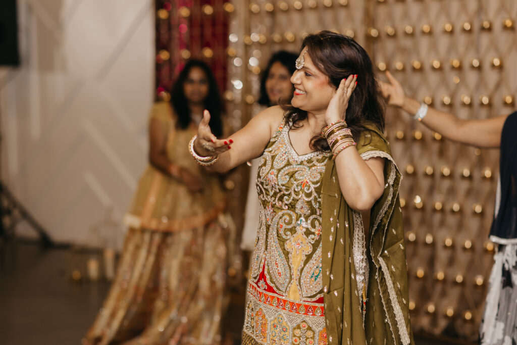 Close-up of a woman in a green and gold outfit joyfully dancing with hand near her ear during a sangeet, surrounded by friends in the background