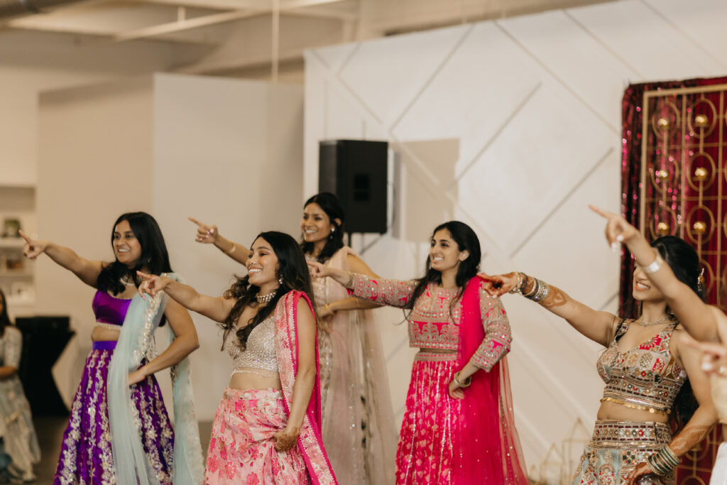 Group of women dressed in vibrant pinks and purples performing a coordinated dance routine at a sangeet celebration inside a modern venue