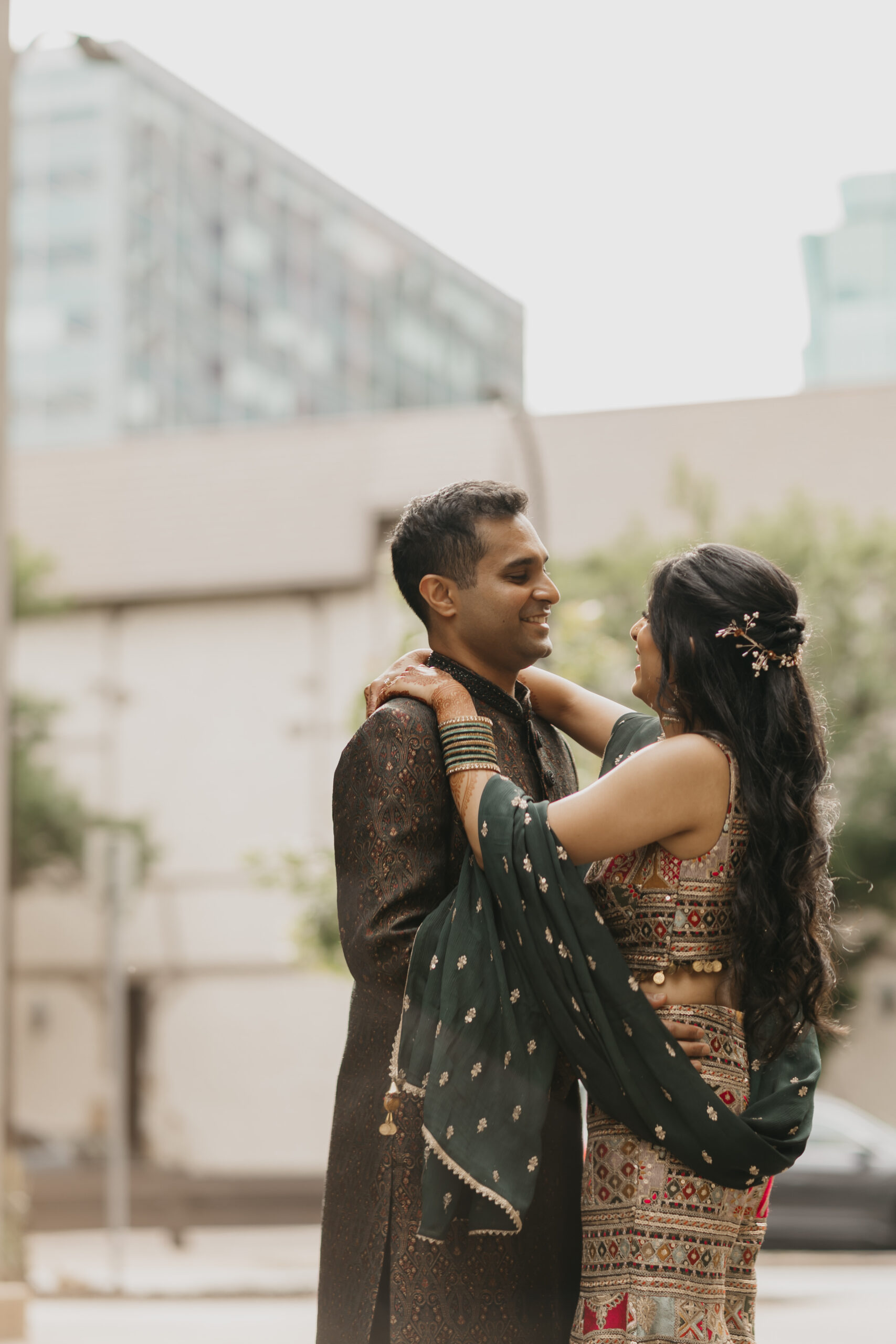 Indian couple smiling at each other in downtown Minneapolis during their sangeet, wearing traditional embroidered outfits in deep green and gold tones