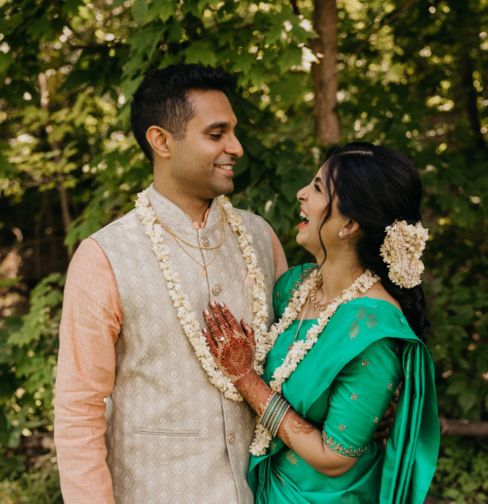 Groom twirling his bride in a lush green outdoor setting during a traditional Indian wedding ceremony; the bride wears a vibrant teal saree with gold details, adorned with floral garlands and mehndi-covered hands.