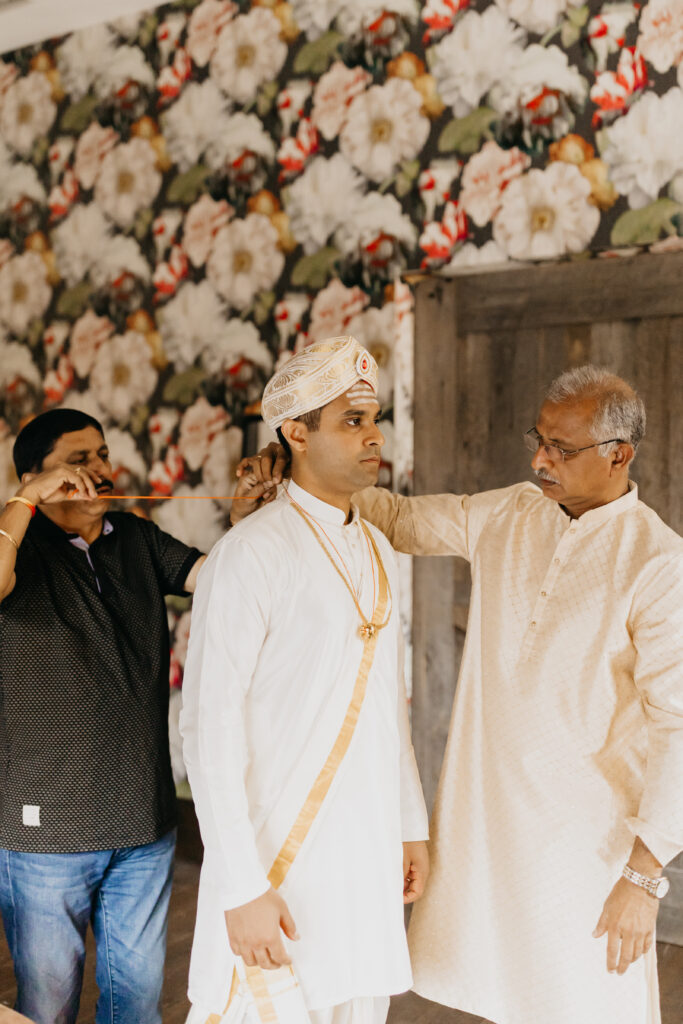 Groom in cream and gold attire with pheta turban, supported by father before wedding ceremony