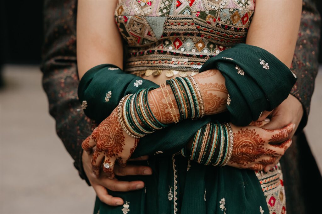 Close-up of the bride’s henna-covered hands resting on her waist, showcasing green and gold bangles and intricate embroidery on her lehenga