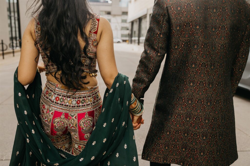 Rear view of the bride holding hands with the groom while walking outside, highlighting the details of her open-back lehenga and flowing dupatta