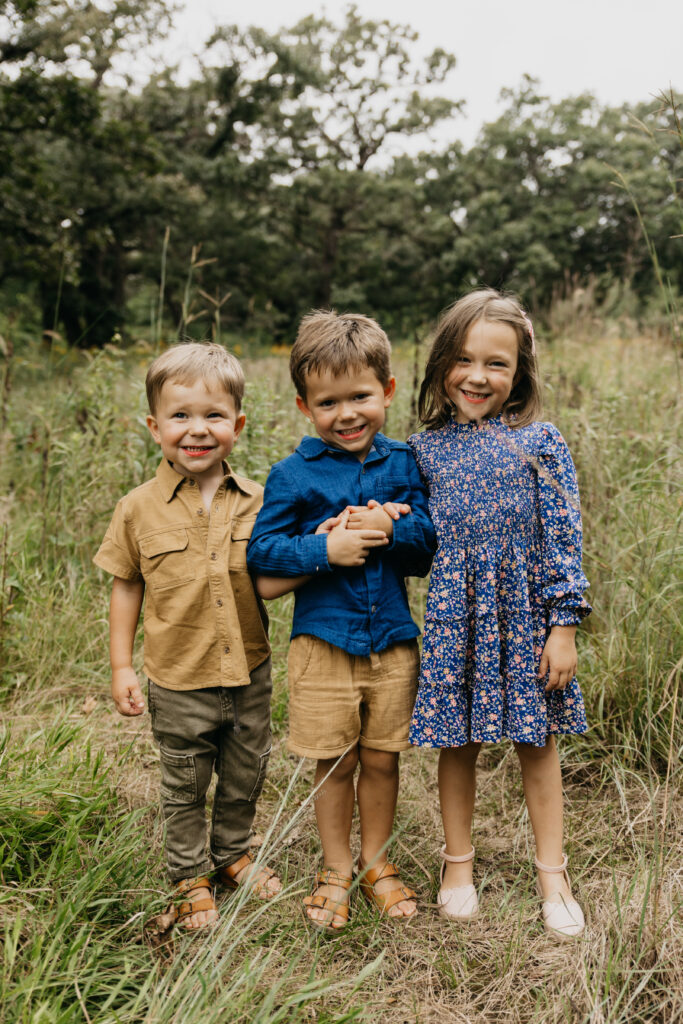 Three siblings standing close together and smiling in a tall grassy field during a Minnesota family portrait session.