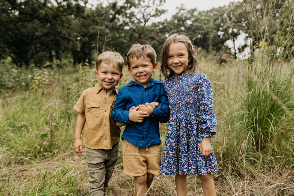Two brothers holding hands and smiling next to their sister in a grassy field during a Minnesota family photo session.