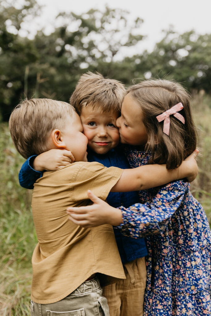  Three young siblings sharing a big hug in a grassy Minnesota field during a family photo session.