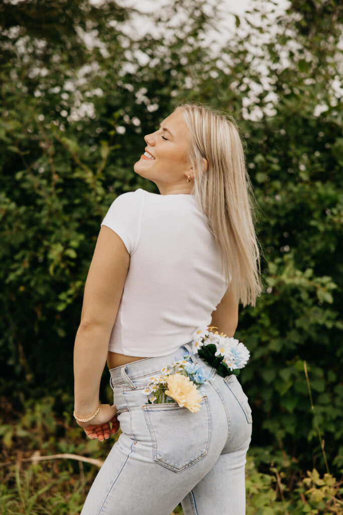 High school senior in a white crop top and light-wash jeans with colorful flowers tucked into her back pocket, smiling during senior photos in Minneapolis by Mycah Bain