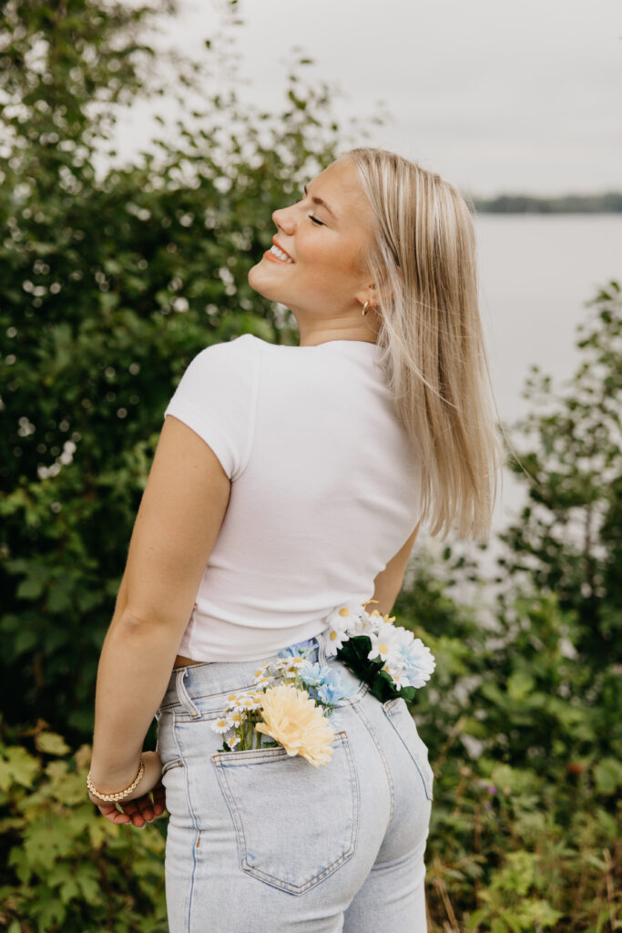 Senior girl smiling with flowers tucked into the back pocket of her jeans, posing lakeside during her Minneapolis senior photo session with Mycah Bain Photography.