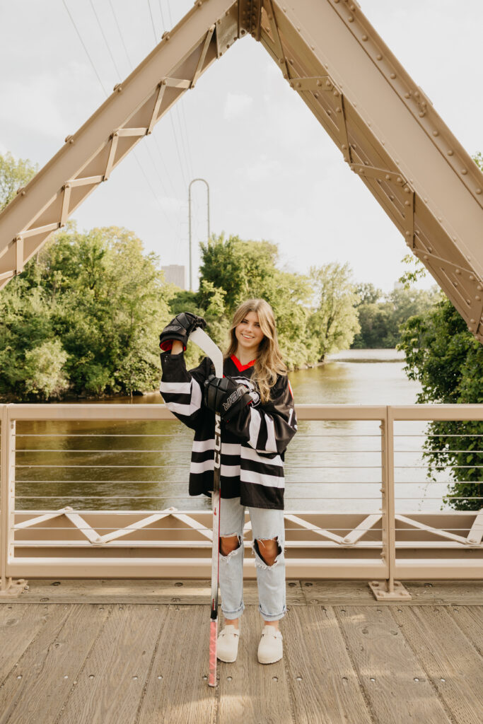 Senior girl in a hockey jersey and gloves holding her stick, standing on a bridge with trees and river in the background during her Minneapolis senior portraits with Mycah Bain Photography.
