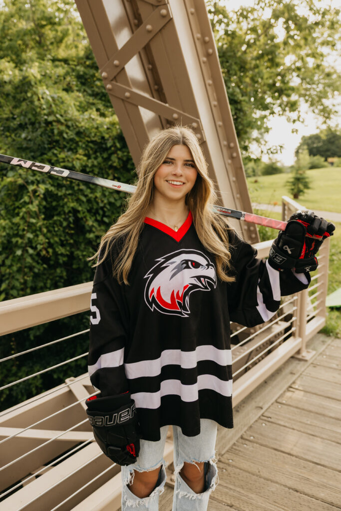 High school senior in ripped jeans and hockey jersey posing with her stick resting on her shoulders, captured outdoors on a bridge by Mycah Bain Photography in Minneapolis.