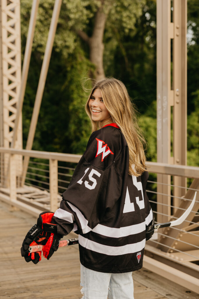 Senior girl smiling while holding a hockey stick and wearing her black and red team jersey, standing on a bridge during her Minneapolis senior photos with Mycah Bain Photography.