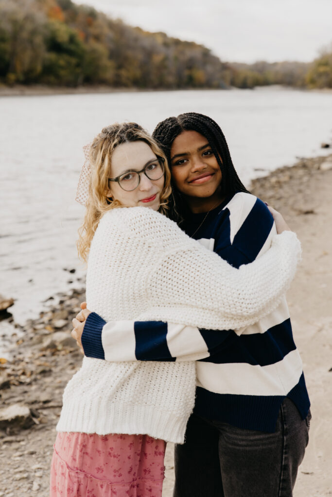 Two sisters hugging and smiling by the riverbank during a fall family portrait session in Minnesota.