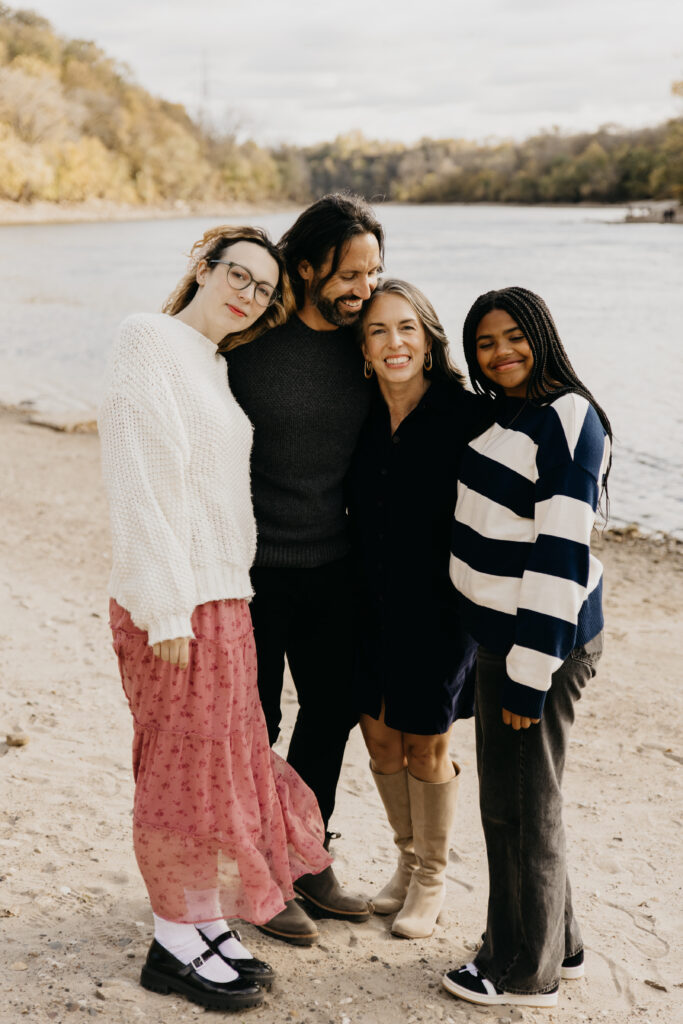 Family of four embracing by the river during a sunny fall day in Minnesota, wearing cozy sweaters for a family photo session.