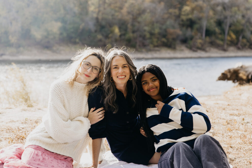 Mother sitting between two daughters on a riverside, all smiling and enjoying a Minnesota fall family photo session.