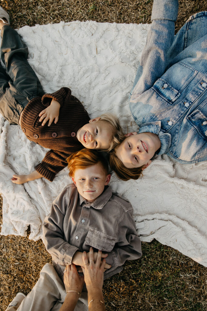 Three siblings lying on a blanket outdoors, smiling and relaxing together during a cozy Minnesota family photo session.