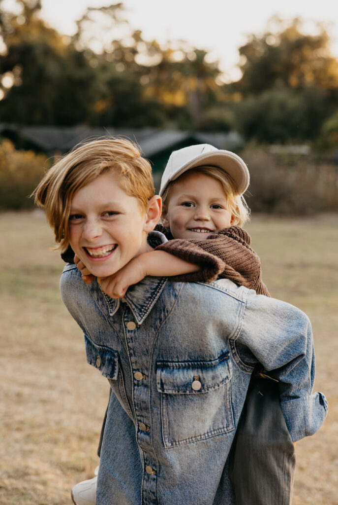 Older brother giving younger sibling a piggyback ride while smiling at the camera during a sunny Minnesota family photo session.