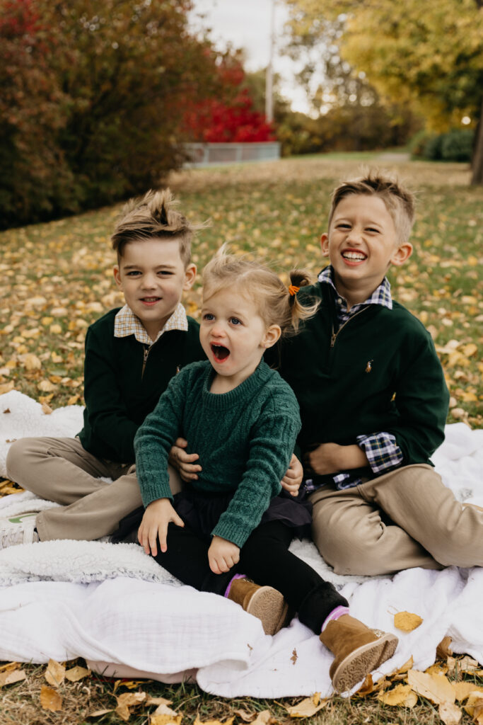  Three siblings sitting on a blanket surrounded by autumn leaves, smiling and laughing during a Minnesota family photo session.