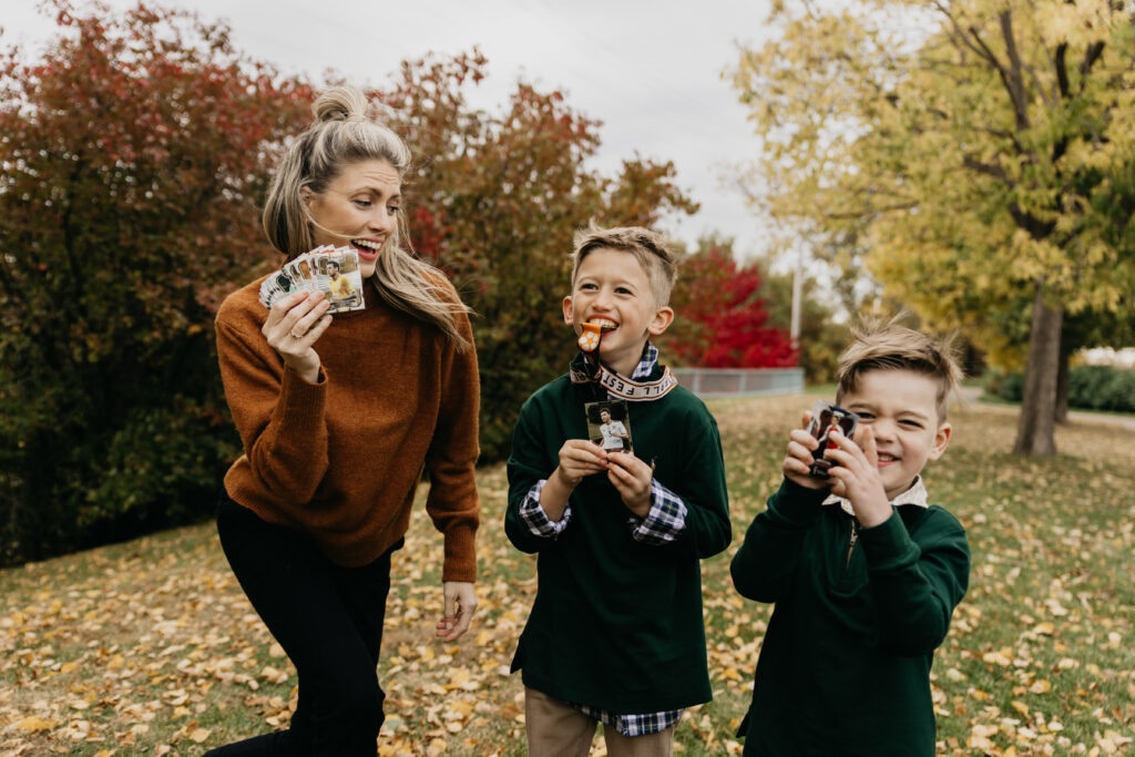 Mother holding holiday photo cards while standing with her two young sons, all smiling and celebrating outdoors in a Minnesota park during fall.