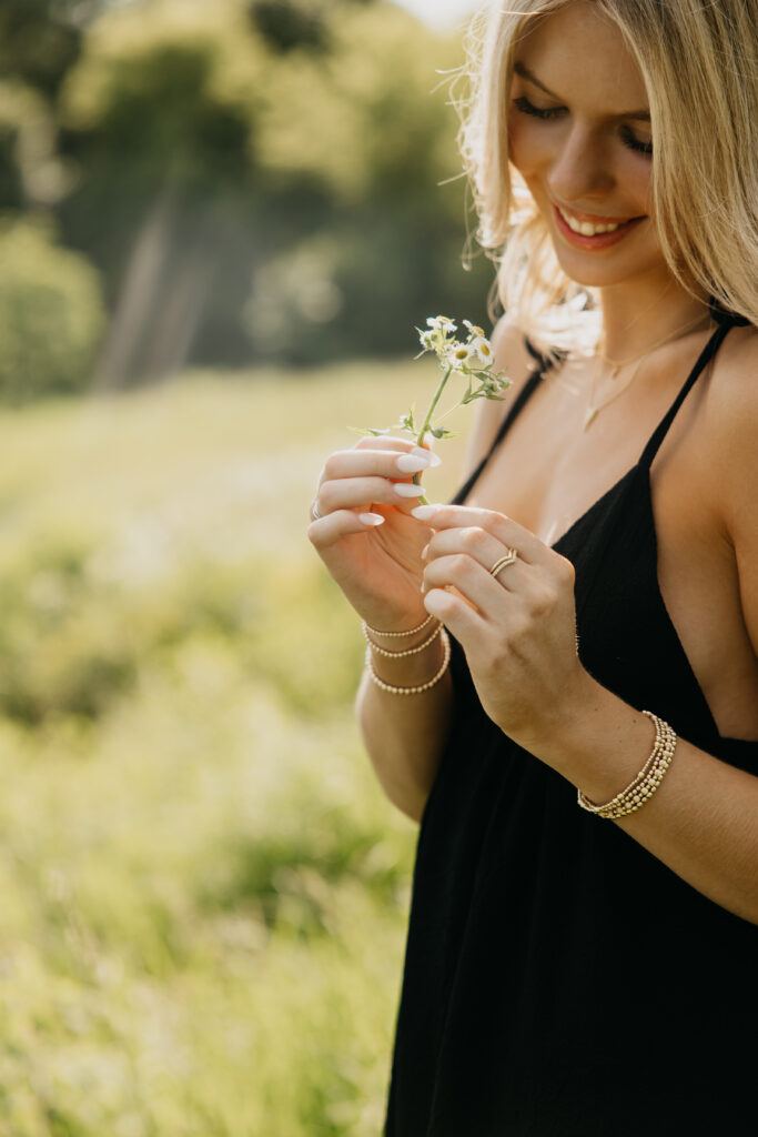 High school senior smiling while looking down at a wildflower, wearing a black dress in a sunlit grassy field, captured by Mycah Bain Photography in Minneapolis.