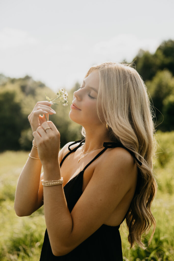 Senior girl in a black dress holding a small wildflower with eyes closed, standing in a sunny green field during golden hour senior portraits by Mycah Bain Photography.