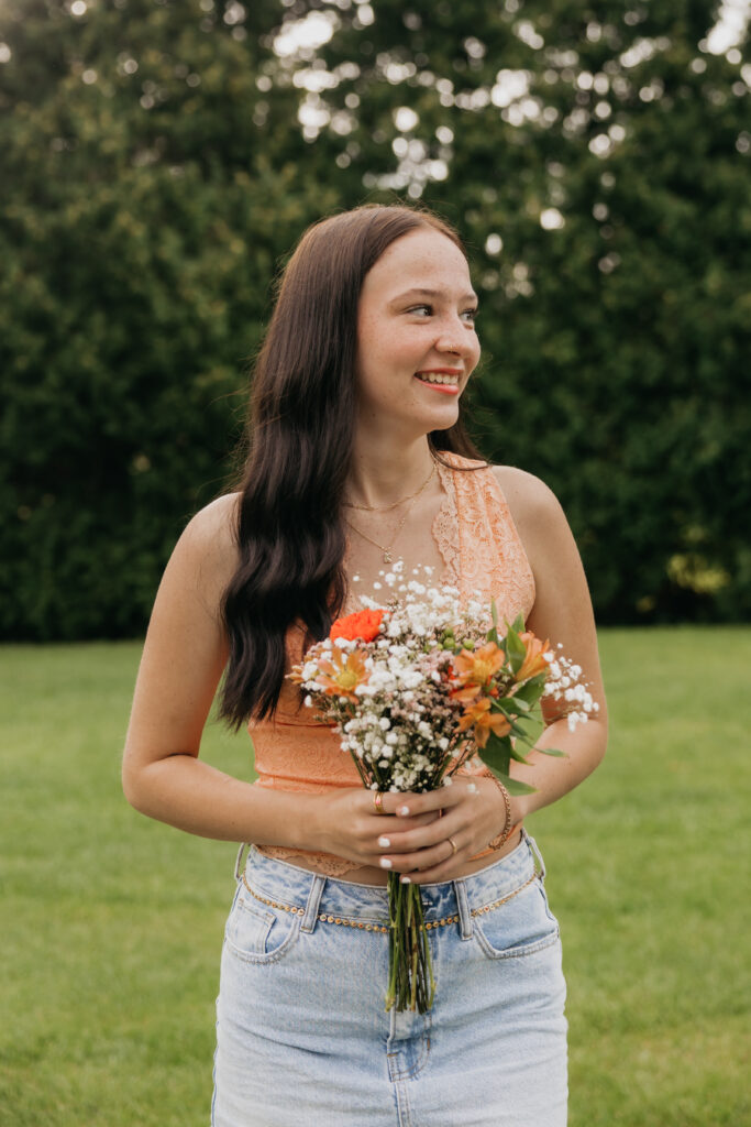 High school senior in an orange lace top and denim shorts holding a colorful flower bouquet, smiling outdoors in a grassy field during her Minneapolis senior photo session with Mycah Bain Photography.